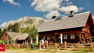 Two people stand in front of a wooden house with solar panels on the roof, surrounded by a wooden fence and a mountain in the background.