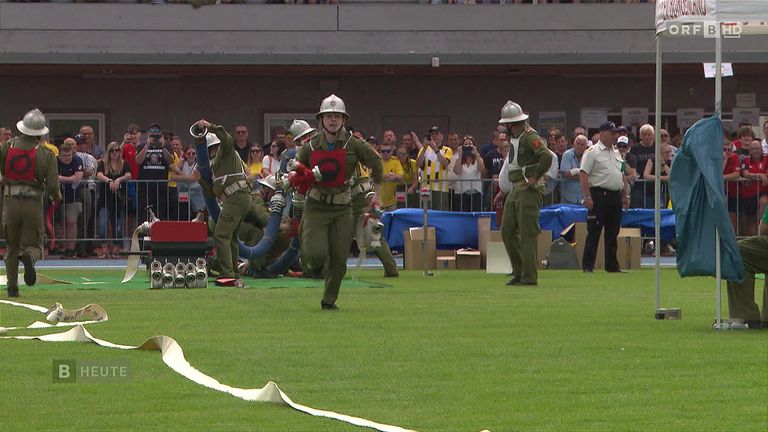 A group of people in military uniforms are performing a drill on a field. Spectators watch from behind a fence.