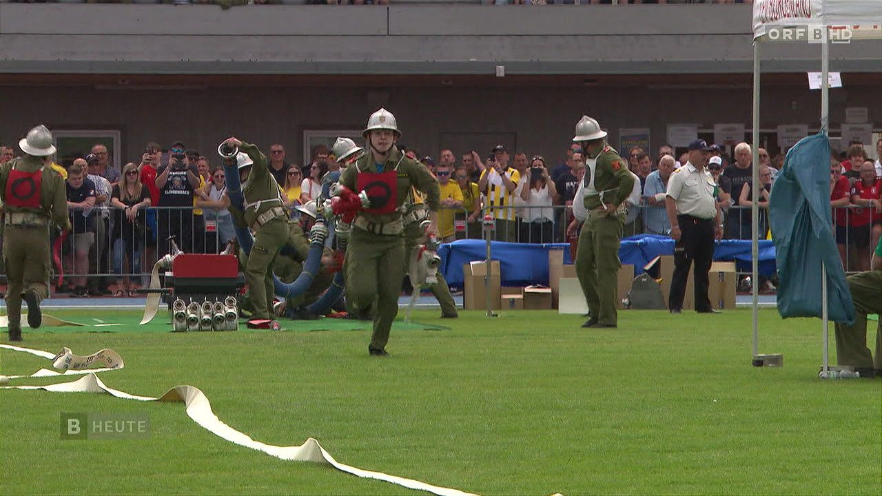 Eine Gruppe von Menschen in Militäruniformen führt eine Übung auf einem Feld durch. Zuschauer beobachten hinter einem Zaun.