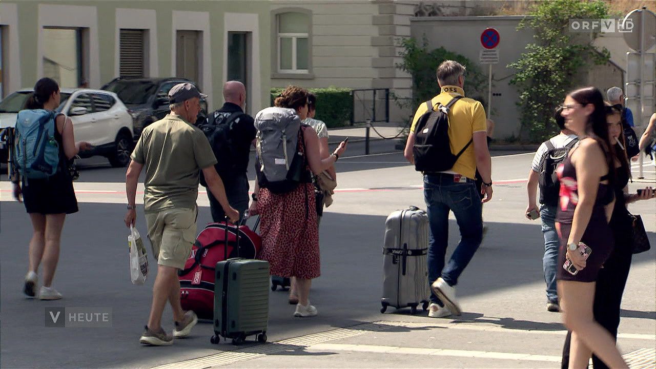 A group of people are walking on a city street with luggage. One person is using a smartphone. Buildings and a car are in the background.