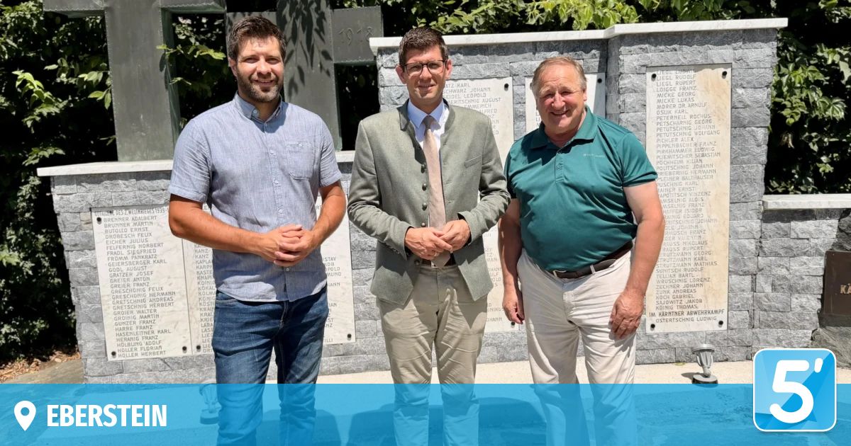 Three men stand together in front of a memorial wall with plaques, smiling for a photo. The man on the left wears a light blue shirt and jeans, the middle man a light green blazer and khaki pants, and the man on the right a dark green polo shirt and khaki pants.