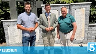 Drei Männer stehen lächelnd vor einer Gedenkwand mit Tafeln und lassen sich fotografieren. Der Mann links trägt ein hellblaues Hemd und Jeans, der Mann in der Mitte einen hellgrünen Blazer und Khakihosen, und der Mann rechts ein dunkelgrünes Poloshirt und Khakihosen.