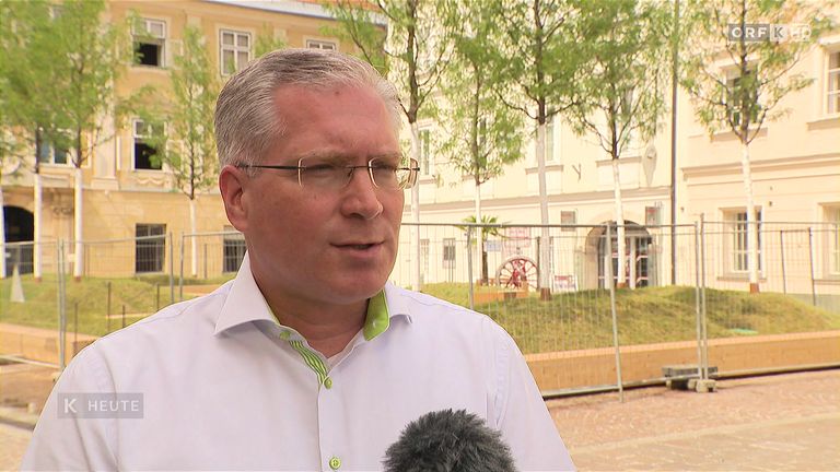 A man in a white shirt with green trim speaks into a microphone, with a building and trees in the background.
