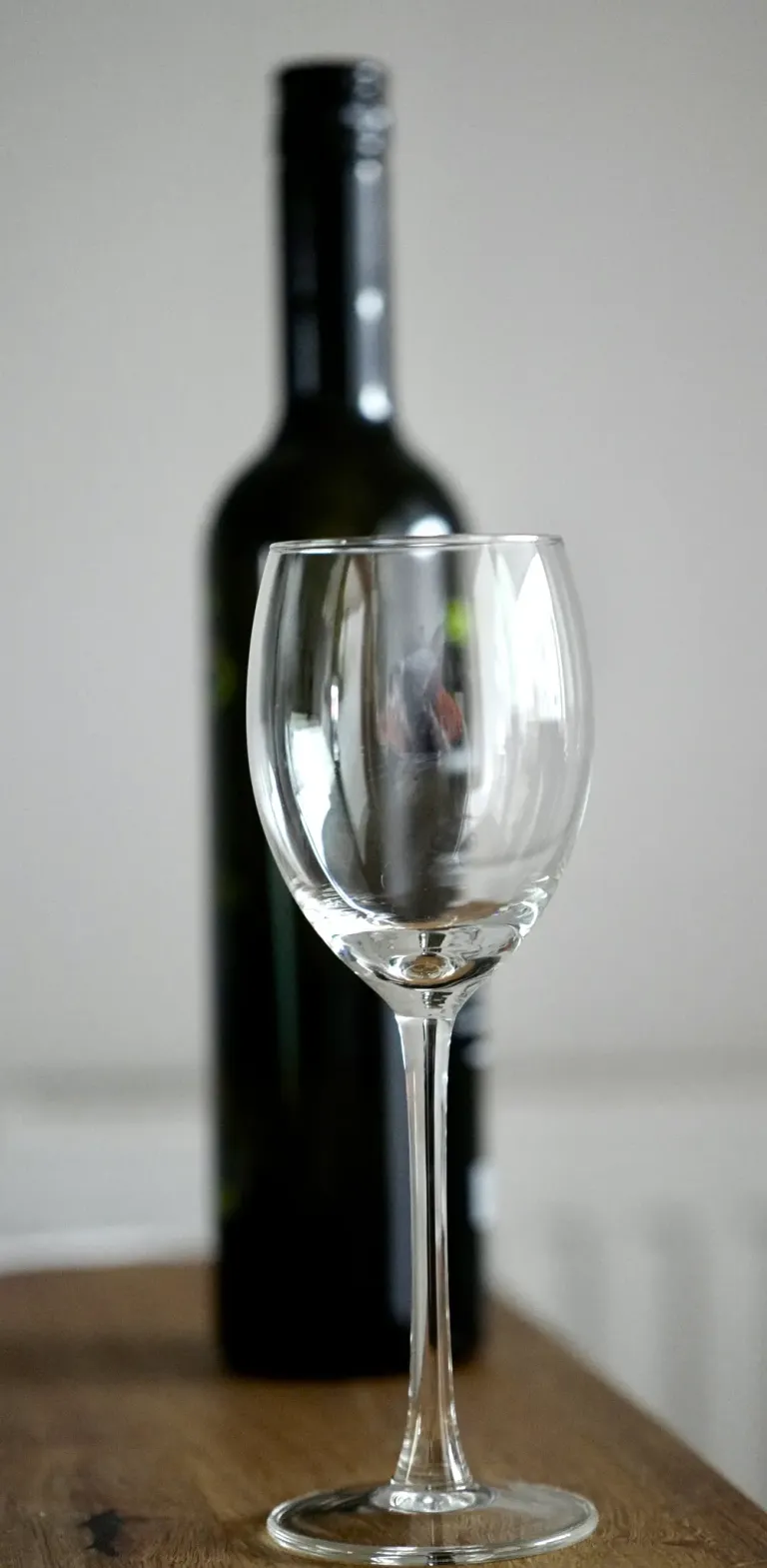 A clear wine glass is positioned in front of a dark green wine bottle, both on a white background.