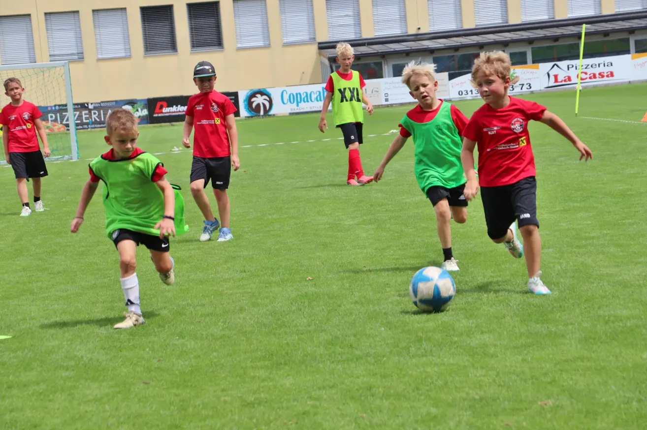 A group of young boys in green and red soccer jerseys are playing soccer on a field. One boy is running towards the ball.