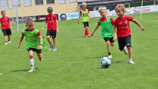 A group of young boys in green and red soccer jerseys are playing soccer on a field. One boy is running towards the ball.