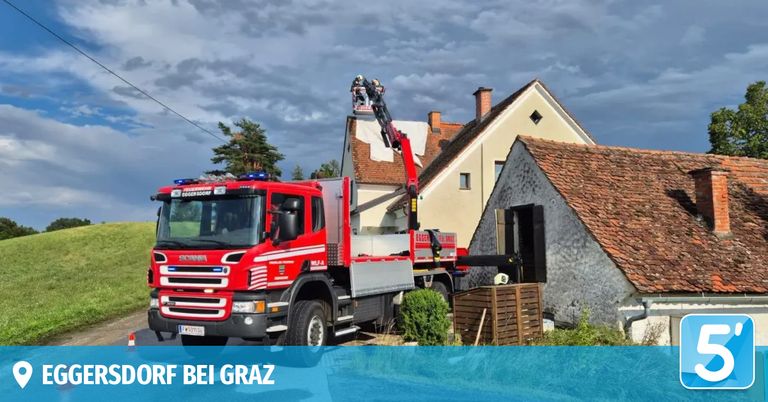 Ein roter Feuerwehrwagen mit einem Kran steht neben einem Haus mit einem Ziegeldach. Zwei Feuerwehrleute sind auf dem Kran, und der Wagen hat das Wort Scania auf seiner Front.