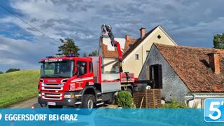 A red fire truck with a crane is positioned next to a house with a tiled roof. Two firefighters are on the crane, and the truck has the word Scania on its front.