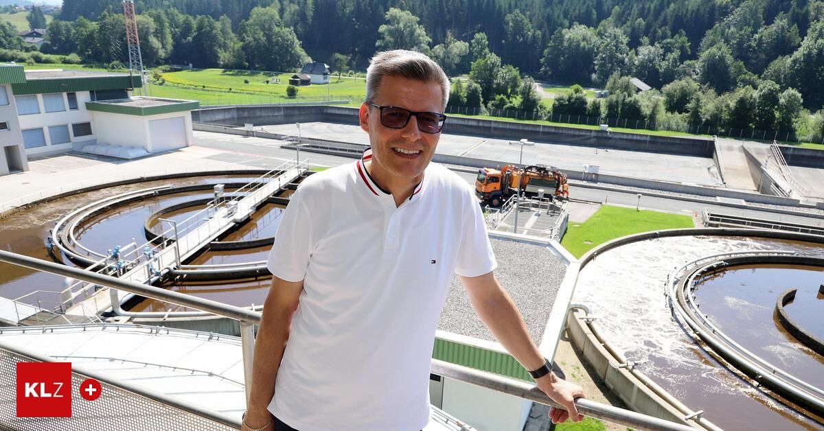 A man in a white shirt stands on a balcony overlooking a wastewater treatment plant. He is smiling and looking at the camera. Behind him, a truck is parked on the road.
