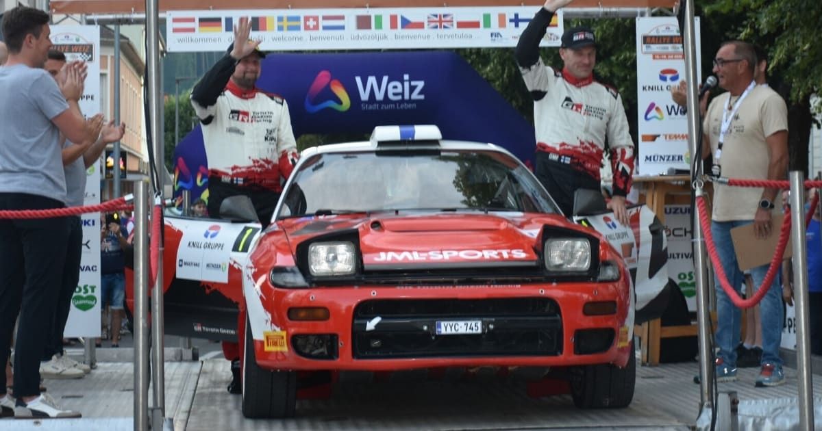 Two men stand beside a red rally car with the license plate YYC-745, sponsored by JML Sports. They are waving to the crowd, and the car is on a platform. Behind them is a banner with the text 'Weiz' and various flags.