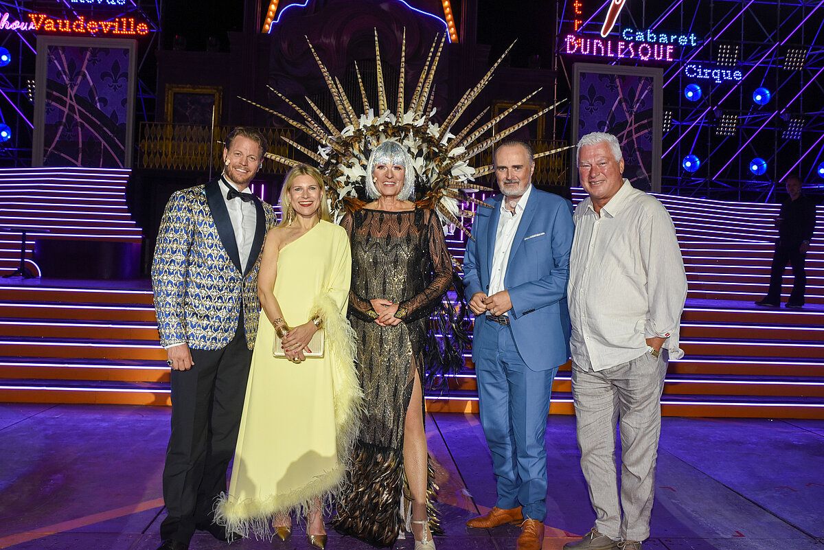 Fünf Personen stehen auf einer Bühne und lächeln für ein Foto. Eine Frau mit einer Federhaube steht in der Mitte. Hinter ihnen befindet sich ein Neonschild, das 'Burlesue Cabaret' besagt.