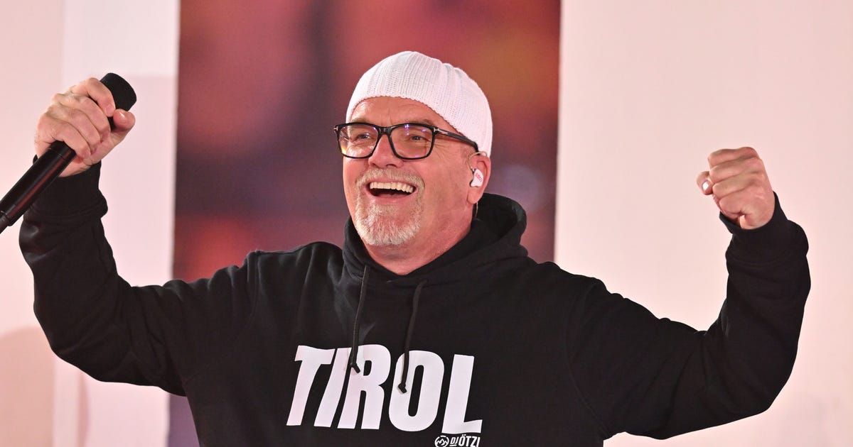 Ein Mann mit einer weißen Mütze und Brille, lächelt und posiert mit weit ausgestreckten Armen. Er trägt eine schwarze Kapuzenjacke mit dem Wort 'TIRO' darauf.