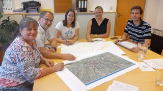 Five people sit around a table with a large map, smiling and looking at it. One person holds a pen and points to the map.