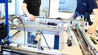 Two individuals are working on a complex machine in a factory setting. The machine has numerous wires and components, and the person on the left is interacting with a control panel.