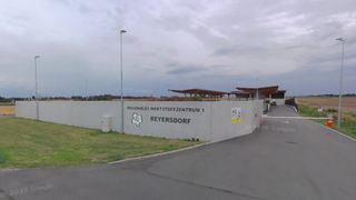 A large white wall with a logo and text 'REGIONALES WERTSTOFFZENTRUM 1 REYERSDORF'. A street light is on the left side. A road with a car is on the right.
