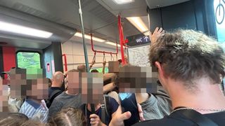 A crowded train with passengers holding onto poles and standing. A person in the foreground is holding a phone.