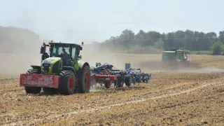 A green tractor with a plow attachment is working in a field, with another tractor visible in the background. The field is dry and dusty.