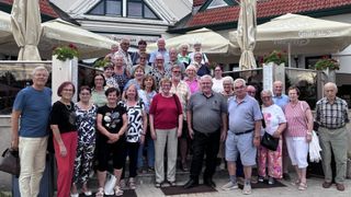 A group of elderly people are standing together outside a restaurant, smiling for a photo. They are wearing casual clothes and some are wearing glasses. Behind them are umbrellas and a building with a sign.