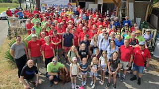 A large group of people in red shirts, some wearing hats, glasses, and sneakers, are posing for a photo in an outdoor area. A man in a green shirt is kneeling with a dog. In the background, there is a banner with the text 'Erich Hoejschi'.