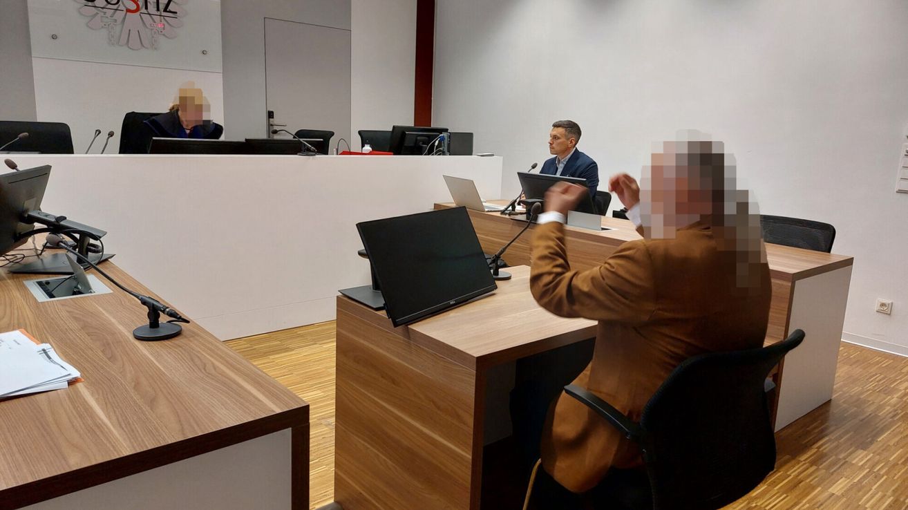 Ein Mann im Anzug spricht in ein Mikrofon an einem Schreibtisch in einem Konferenzraum. Ein anderer Person sitzt vor ihm.