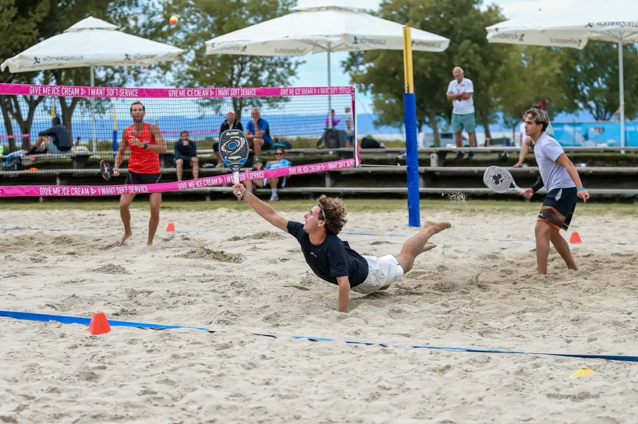 Ein Mann taucht auf dem Sand, um einen Beachvolleyball zu schlagen, während ein anderer Mann hinter dem Netz zusieht.