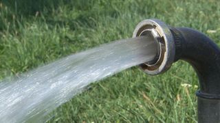A close-up view of a water hose spraying water onto a grassy field. The hose is made of metal and has a threaded end.