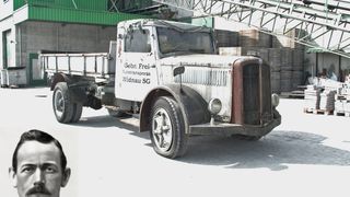 An old Sauer-Diesel truck with the text 'Gebr. Frei-Autotransporte Widnau SG' parked in front of a warehouse with stacks of wooden pallets.