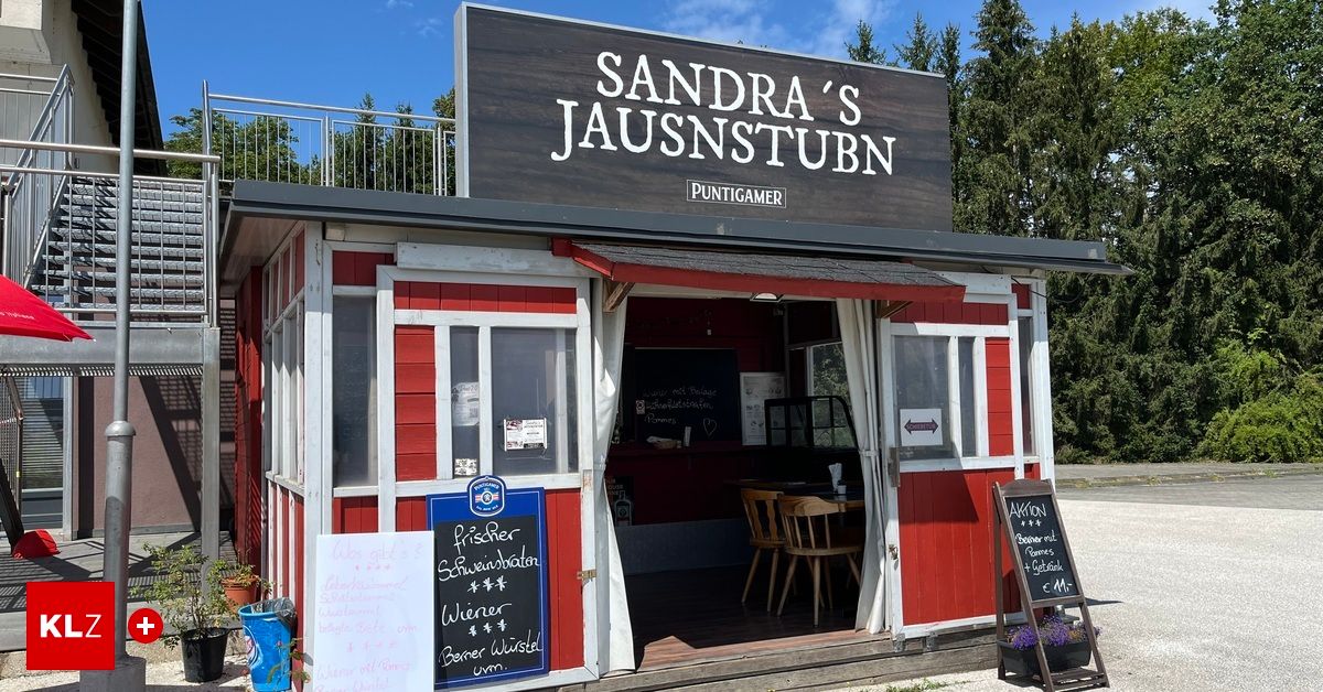 Ein kleines rotes und weißes Gebäude mit einem Schild, das 'Sandra's Jausnstubn' besagt. Im Inneren befinden sich Holzstühle und ein Tisch. Ein Kreidetafelmenü ist auf der linken Seite sichtbar, und eine Topfpflanze mit lila Blumen steht rechts.