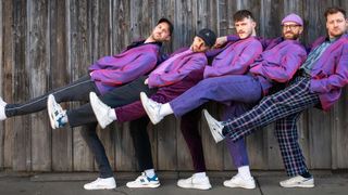 Four men in matching purple suits pose against a wooden wall, with one man sitting on the shoulders of another, creating a dynamic and playful scene.