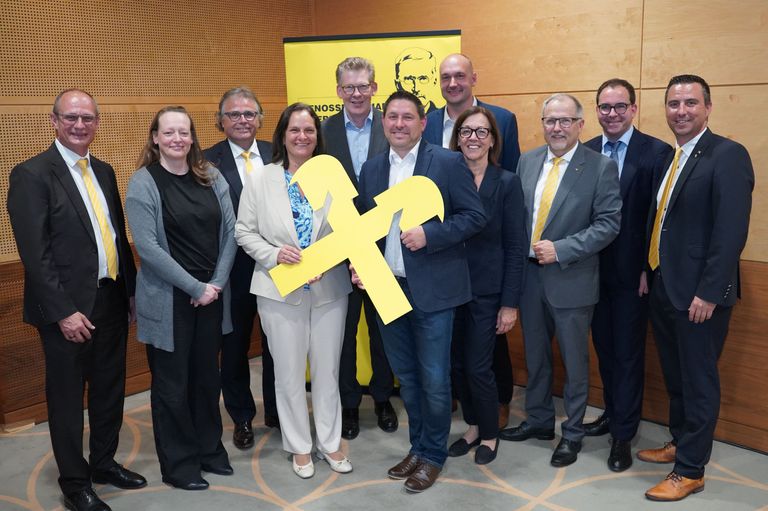 Eine Gruppe von Fachleuten in formeller Kleidung, die zusammenstehen, einer hält einen großen gelben Buchstaben F, mit einem gelben Banner im Hintergrund.