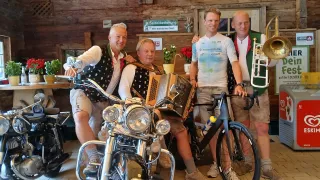 Four men in traditional clothing pose with a motorcycle and a bicycle, one man plays an accordion.
