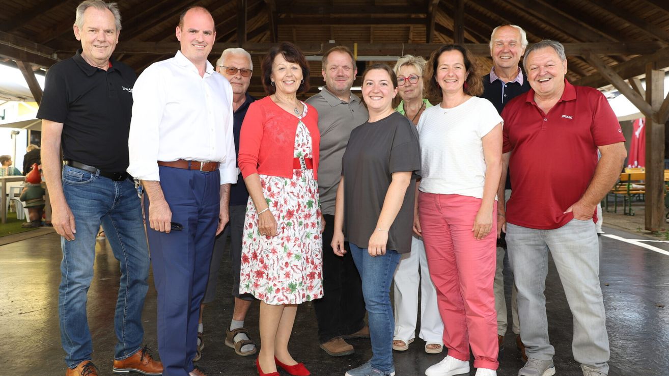 A group of people, including men and women, are standing together in a room, smiling for a photograph. They are dressed in casual clothing.