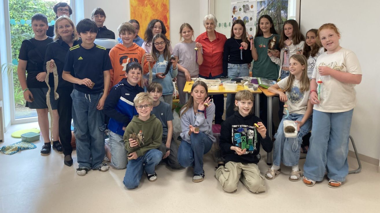 A group of children and adults are posing for a photo in a classroom. Some children are holding small animals, and a woman in a red shirt is standing behind them.