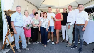 A group of people, including men and women, are standing together in a tent. Some are holding wine glasses, and one person is holding a bouquet of flowers.