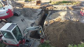 A construction site with a large trench being dug by a white excavator. A yellow excavator is parked in the background.