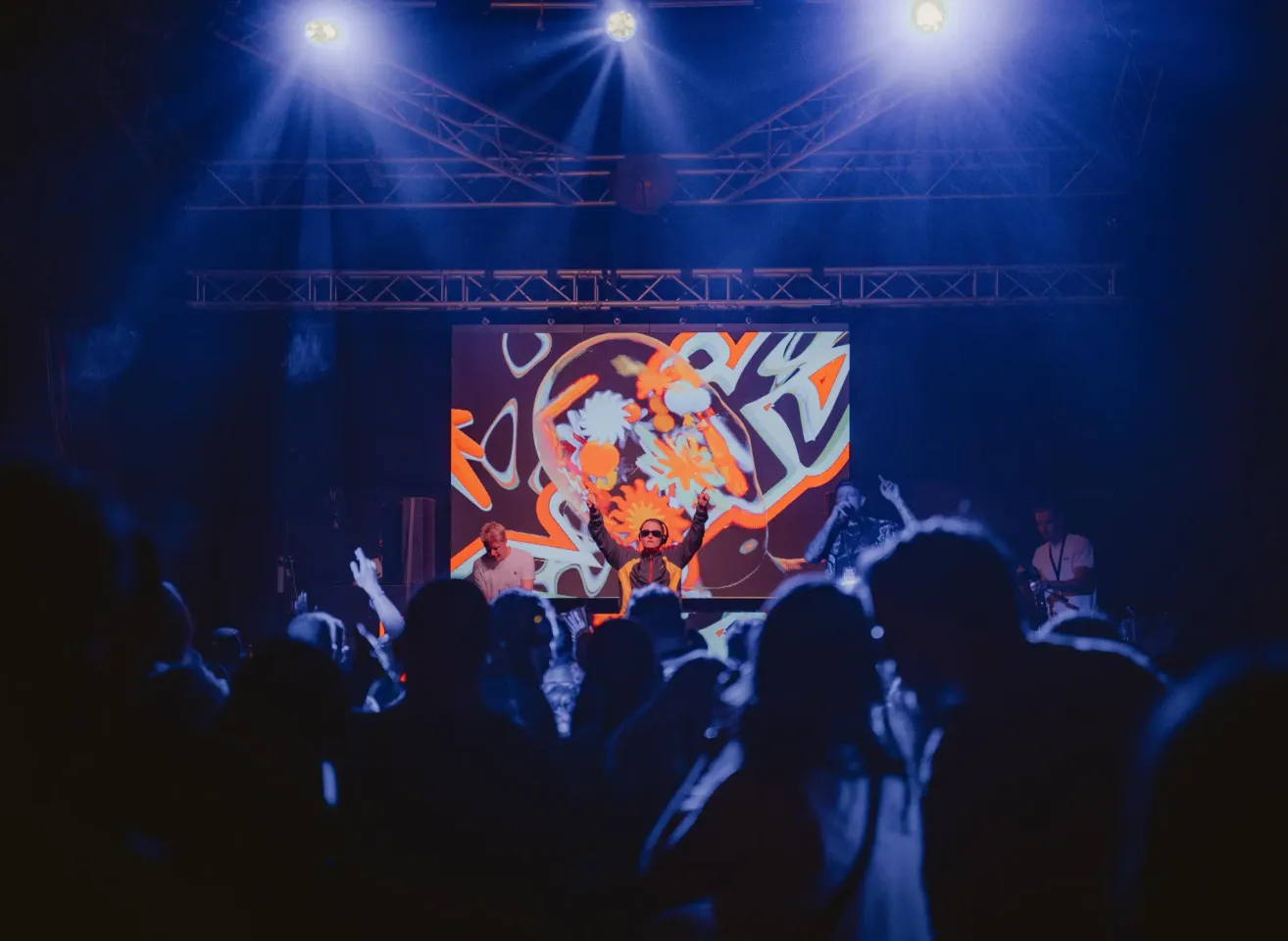 Ein DJ tritt auf der Bühne auf, mit einer großen Leinwand, die abstrakte Kunst hinter ihm zeigt, unter blauen Bühnenlichtern, während eine Menge zuschaut.
