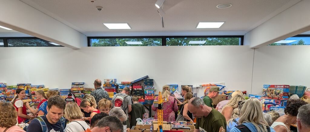 A room filled with people browsing through a variety of toys and games, with a large window showing trees outside.