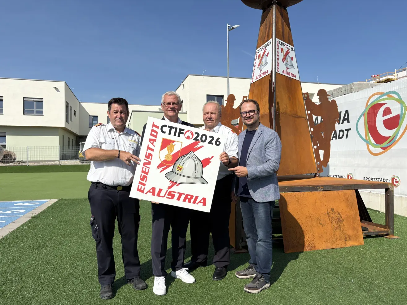 Four men stand on a green field, holding a sign that reads 'CTIFO 2026 Eisenstadt Austria'. Behind them is a large sculpture.