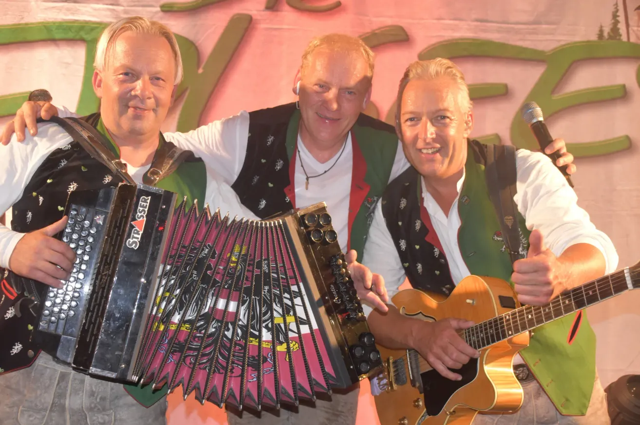 Three men dressed in traditional attire, one playing an accordion, another playing a guitar, and the third smiling at the camera. They are standing in front of a pink backdrop.