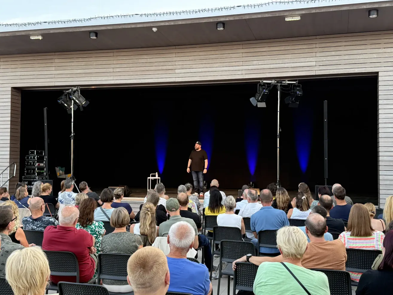 A man stands on stage in front of a seated audience, illuminated by blue stage lights. The audience is diverse in age and attire, attentively watching the speaker.