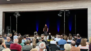 A man stands on stage in front of a seated audience, illuminated by blue stage lights. The audience is diverse in age and attire, attentively watching the speaker.