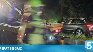 A tow truck with a flatbed is towing a damaged car on a wet road at night. The car is tilted to the side, and the tow truck's lights are on.