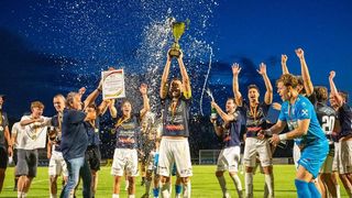A group of soccer players celebrate a victory on a field at night, holding a trophy and champagne. They are surrounded by confetti and cheering fans.
