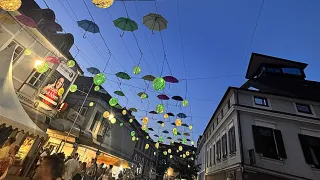 A bustling street at dusk, adorned with colorful umbrellas and lanterns hanging from wires above. People are walking and shopping in the area.