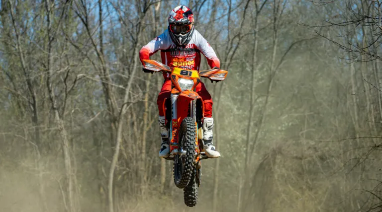 Ein Motocross-Fahrer in voller Ausrüstung, einschließlich Helm und Handschuhen, springt in der Luft auf einer Dirt-Piste mit Bäumen im Hintergrund.