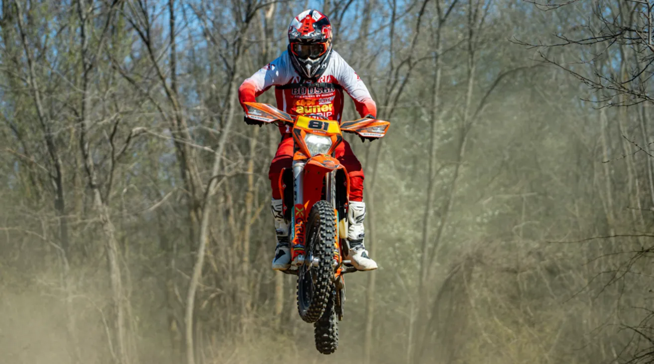 A motocross rider in full gear, including helmet and gloves, jumps mid-air on a dirt track with trees in the background.