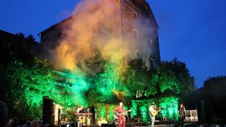 A band performs on a stage at night, with a large building in the background and colorful lights illuminating the scene.