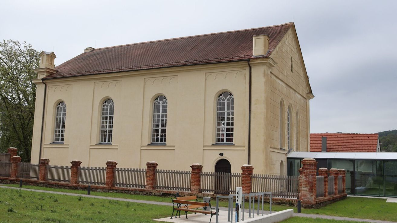 A beige building with a brown roof and arched windows. A wooden bench is in front of it.