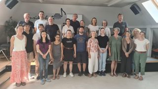 A group of people, both men and women, are standing together in a room, smiling and posing for a photo. They are dressed casually. Behind them, a wall with a musical note design is visible.