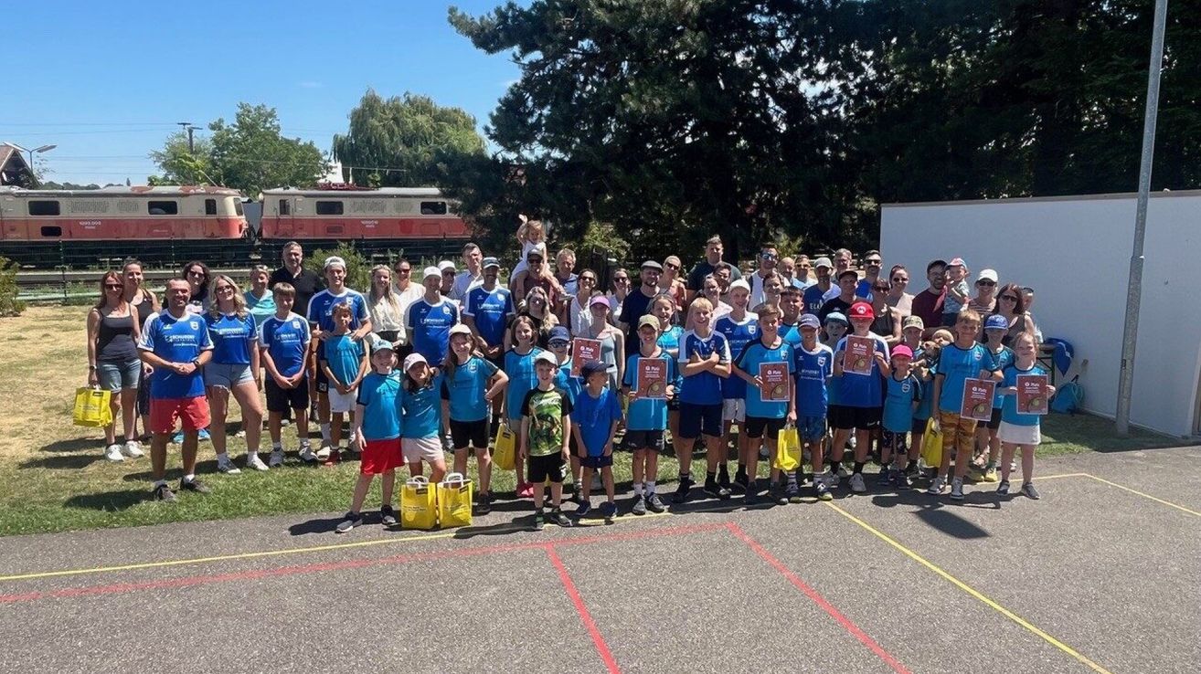 A group of people, including children and adults, are standing in a line on a paved area. They are all wearing blue shirts and holding certificates. Some of them are wearing hats and sneakers. They are probably participating in a sports event. Behind them are trees and a train on the tracks.